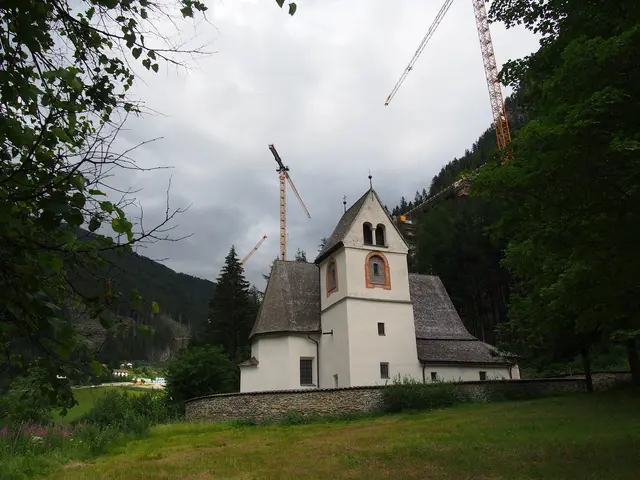 Weißes Kirchengebäude mit spitzem Schindeldach im Tal unter einer großen Beton-Autobahnbrücke und mehreren gelben Kräne.