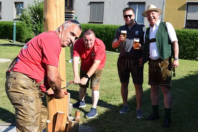 Zwei Männer sägen einen Maibaum um. Zwei Männer stehen mit einem Bier in der Hand daneben