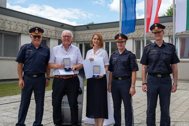 Drei Männer in Polizeiuniform stehen links beziehungsweise rechts von einem Mann und einer Frau, die eine Schachtel mit einer Medaille sowie eine Urkunde in Händen halten. 