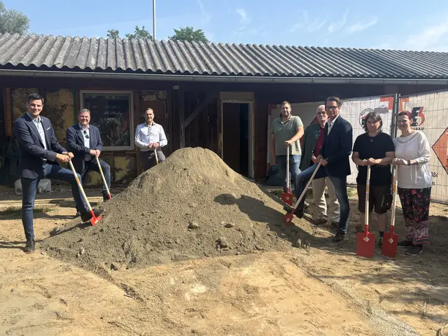 Gruppe von Menschen mit Schaufeln vor Sandhügel und Holzgebäude.