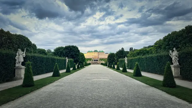 Ein gerader Kiesweg führt zentral durch eine Symmetrie aus hohen, dunkelgrünen Heckenwänden. Am Anfang der Allee stehen links und rechts weiße Barockskulpturen auf Sockeln. Kleine, kegelförmige Bäume säumen den Weg. Am Ende des Weges, in der Ferne, liegt das beleuchtete Obere Belvedere Schloss mit seinem markanten grünen Dach. Der Himmel ist bedeckt mit vielen strukturierten grauen Wolken.