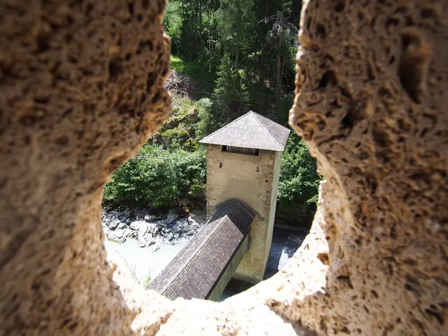 Fotografie aus dem schattigen Inneren von Schloss Sigmundseck. Der Blick führt durch den schmalen Lichtschlitz einer gemauerten Schießscharte hinunter auf den historischen Brückenturm, der mitten im tosenden Inn auf der Holzbrücke steht.
