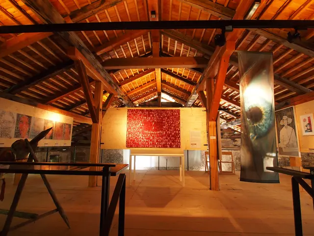 Fotografie aus dem hellen Ausstellungsraum im ausgebauten Dachboden des Klausenturms. Sichtbar sind die mächtigen, historischen Holzbalken der Dachkonstruktion, vor denen moderne Kunstwerke und Gemälde auf Staffeleien präsentiert werden.