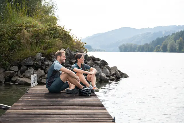 Auf einem Holzsteg an der Donau im Naturhafen Freizell sitzt ein Paar entspannt und genießt ruhige Minuten entlang der Penzensteinrunde des Escape Trails Neustift. | Foto: Tourismusverband Donau Oberösterreich/infilmity