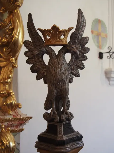 Eine historische dunkelgraue Skulptur eines Doppeladlers mit ausgebreiteten Flügeln und einer goldenen Krone auf den beiden Köpfen, platziert als theologisches Symbol am Hochaltar im Innenraum der Kirche am Lueg