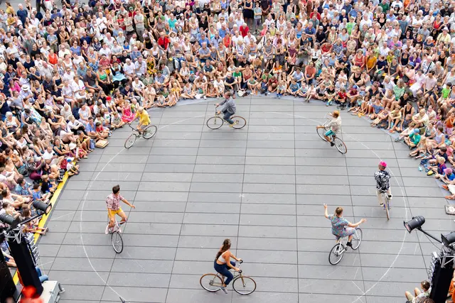 Eine Gruppe an Künstlerinnen und Künstlern fährt im Kreis Fahrrad. Rundherum sitzen und stehen Zuschauerinnen und Zuschauer.