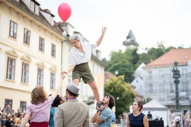 Eine Artistin balanciert auf den Händen anderer Artistinnen und Artisten. Sie bekommt einen Luftballon gereicht.