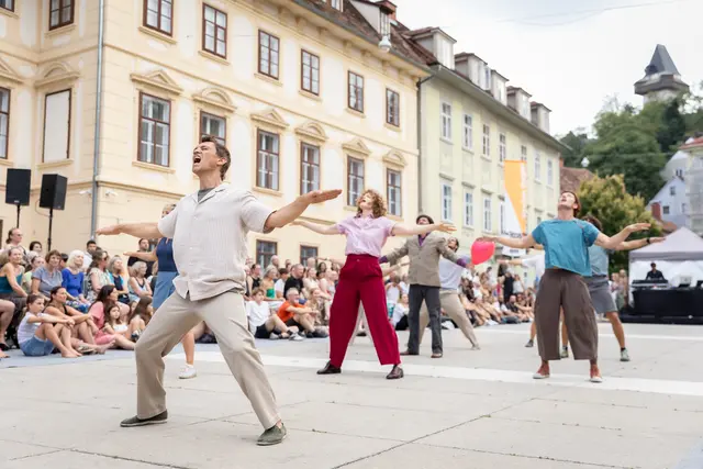 Sieben Personen stehen in einer Pose auf einem Platz und performen vor Publikum.
