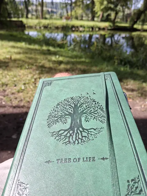 Ein grünes Notizbuch mit dem geprägten Motiv des „Tree of Life“ (Lebensbaums) liegt in der Sonne auf einer Person. Im Hintergrund ist eine unscharfe, idyllische Parklandschaft mit einem Teich zu sehen. Das Motiv des Baumes zeigt auch einige Vögel im Blätterdach. Es symbolisiert die Suche nach neuen Geschichten und literarischen Entdeckungen im Kontext von Kindle Unlimited.