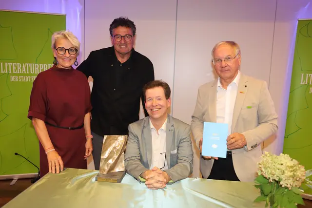 Eine Frau und zwei Männer stehen, davor sitzt ein Mann an einem Tisch 