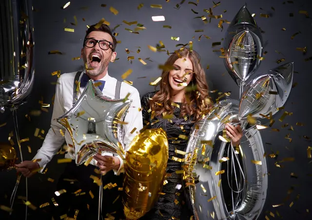 Silvester-Dinner &amp; DJ-Night im Zündwerk Living Room
Auf dem Bild sieht man einen Mann und eine Frau - beide lachen und haben Spaß, halten Luftballone in der Hand und goldenes Konfetti fliegt durch die Luft | Foto: Canva