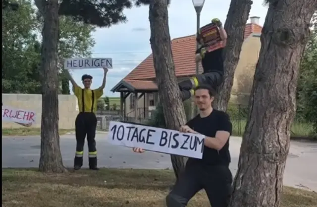 Video-Screenshot: Kameraden mit Schildern zum FF-Heurigen kommen hinter Bäumen hervor