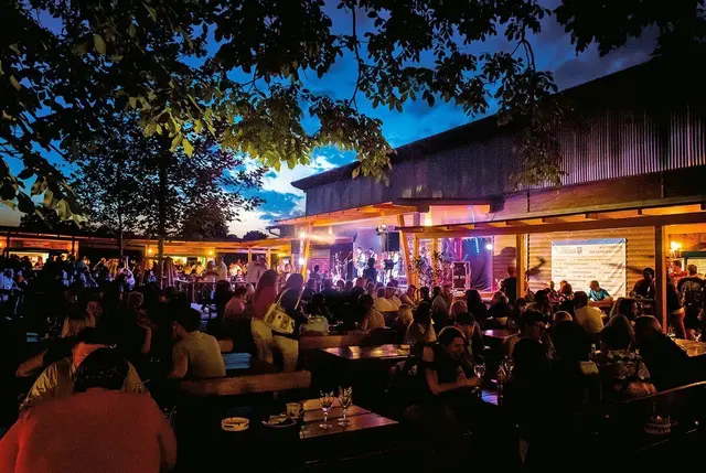 Besucher sitzen bei abendlicher Stimmung bei Biertischen- und Bänken, dahinter Live-Musik