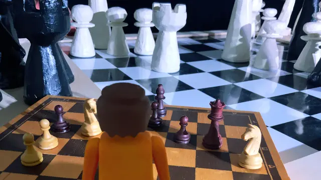 Im Hintergrund ein großes Schachbrett mit Figuren, im Vordergrund ein kleineres Schachbrett mit Figuren, beides aus Holz, davor steht eine Playmobil-Figur, den Schachfeldern zugewandt, de*r*m Betrachter*in den Rücken zugewandt.