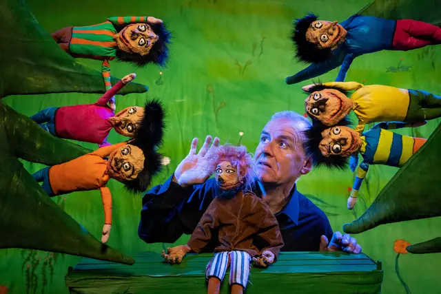 Puppenspieler mit sechs bunten Handpuppen mit schwarzen Haaren und großen Augen auf grünem Bühnenhintergrund.