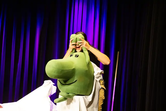 Eine lustige grüne Figur sitzt auf einem weißen Laken, dahinter steht eine Schauspielerin, lachend. Die Schauspielerin hält sich die Hände der grünen Figur vor die Augen, man sieht von ihrem Gesicht nur den lachenden Mund.