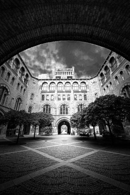 Ein Hof im Wiener Arsenal zeigt sich in einem eindrucksvollen Schwarz-Weiß-Foto. Die tiefstehende Herbstsonne wirft harte Schatten und betont die markanten Kontraste zwischen Licht und Dunkelheit. Die groben Mauern der historischen Gebäude wirken in dem klaren, scharfen Licht fast monumental. Das Spiel von Licht und Schatten verleiht dem Hof eine fast zeitlose Atmosphäre, als ob die Geschichte des Ortes in den Strukturen eingefroren wäre.
