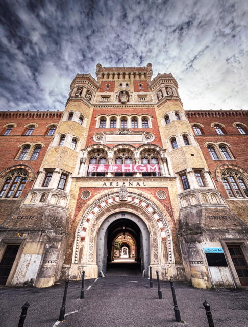 Der imposante Eingangsbereich des Arsenals in Wien ist ein wahres Meisterwerk historistischer Architektur. Mit seinen detailreichen Fassaden, dem markanten Rundbogenportal und den kunstvollen Türmen erzählt dieses Bauwerk Geschichte in Stein. Die dramatische Perspektive, verstärkt durch den weiten Himmel darüber, macht deutlich, wie lohnend die fotografische Auseinandersetzung mit solchen historischen Bauwerken sein kann. Wer Architektur liebt, findet hier unzählige Motive – von der Gesamtansicht bis hin zu ornamentalen Details. Lass dich inspirieren, die Kamera oder das Smartphone zur Hand zu nehmen und selbst auf Entdeckungsreise durch die architektonischen Schätze deiner Stadt zu gehen!