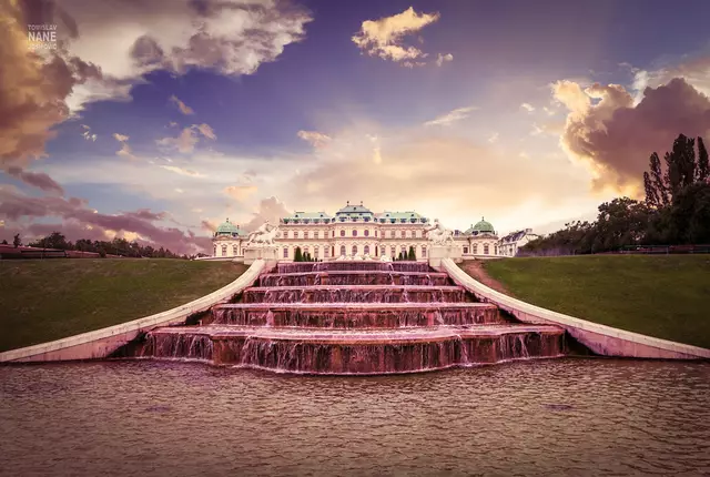Wie doch die Zeit vergeht — still und unaufhaltsam fließt sie dahin, gleich dem Wasser, das in Kaskaden über die kunstvoll angelegten Stufen dieses Brunnens rinnt. Das majestätische Gebäude im Hintergrund, das Belvedere in Wien, hat mehr gesehen, als Worte je beschreiben könnten. Seine Mauern tragen das Flüstern vergangener Jahrhunderte, seine Fenster spiegeln nicht nur das Licht des Sonnenuntergangs, sondern auch das Echo unzähliger Geschichten.

Hier schritten einst Fürsten und Künstler, Liebende und Revolutionäre. Es war Bühne glanzvoller Empfänge, stiller Spaziergänge, und wohl auch Zeuge leiser Tränen. Der Himmel über ihm hat sich in tausend Farben gezeigt – so wie in diesem Moment, wenn der Tag zur Dämmerung wird und alles in ein magisches Licht taucht.

Ein Ort wie dieser erinnert uns daran, dass Geschichte nicht nur in Büchern lebt, sondern in Steinen, in Wasser, im Wind, der durch die Alleen streicht. Und so bleibt das Belvedere nicht bloß ein Schloss, sondern ein Gedächtnis – still, würdevoll, und doch lebendig in jeder Seele, die verweilt und lauscht.

