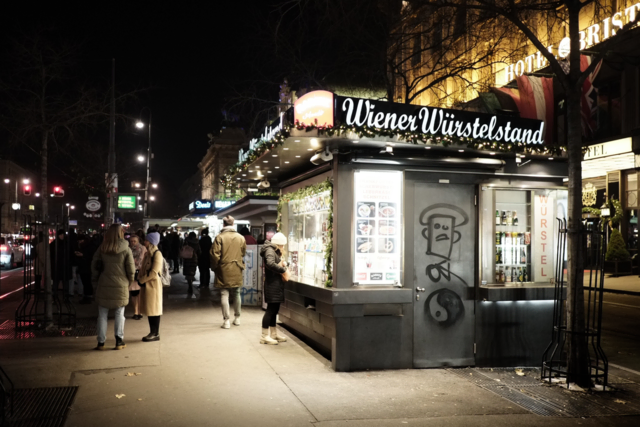 Wien, wenn die Lichter blühen. Am Würstelstand zischt das Fett, der Duft zieht in die kalte Nachtluft. Eine Frau mit weißer Haube kramt in ihrer Tasche, sucht nach Kleingeld, nach einem Moment Ruhe mitten im Trubel.
Die Straßenbahn kündigt sich mit einem metallischen Raunen an. Menschen warten oder eilen vorbei – in die Passage, nach Hause oder tiefer hinein ins nächtliche Wien. Der Stand wird zum Treffpunkt, zur Bühne für kleine Dramen des Alltags.
Hier riecht es nach Würstel, nach Stadtluft, nach Leben. Die Stadt wird langsamer, aber sie schläft nie. Und in diesem Licht, in diesem warmen Schimmer erkennt man Wien, wie es wirklich ist: geschäftig, menschlich, ein wenig melancholisch – und immer hungrig auf mehr.

