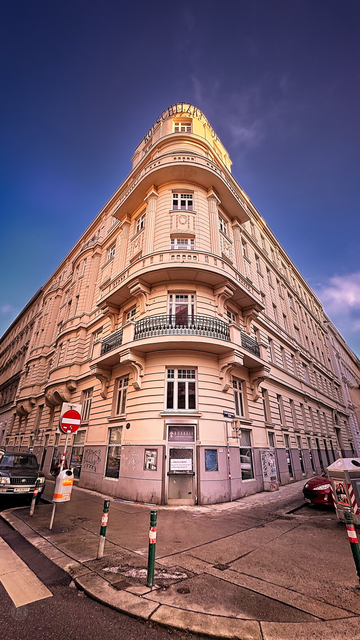 Wer an der Ecke Graf-Starhemberg-Gasse 39 und Kolschitzkygasse im 4. Bezirk hochblickt, entdeckt ein architektonisches Prachtstück, das weit mehr als nur Fassade zu bieten hat: den Kolschitzkyhof.

Ein Denkmal für den „Kaffee-Helden“

Der Name des Hofes ist kein Zufall. Er erinnert an Georg Franz Kolschitzky, der Legende nach der Mann, der den Wienern nach der Belagerung von 1683 das Kaffeetrinken schmackhaft machte. Auch wenn Historiker heute wissen, dass die Geschichte etwas komplexer war, bleibt sein Name untrennbar mit der Wiener Kaffeehauskultur verbunden. Gekrönt wird das Gebäude von der weithin sichtbaren Aufschrift „Kolschitzkyhof“.

Neoklassizismus trifft Dynamik

Erbaut wurde das Eckhaus im Jahr 1911 vom Architekten Ignaz Reiser. Mit seinem neoklassizistischen Dekor und der markanten, abgerundeten Ecke ist es ein Musterbeispiel für die repräsentative Mietshaus-Architektur der späten Kaiserzeit.

Die fotografische Perspektive

In dieser Aufnahme habe ich bewusst das Weitwinkelobjektiv eingesetzt, um die monumentale Wirkung des Gebäudes zu unterstreichen:
Der „Schiffsbug“-Effekt: Durch den tiefen Standpunkt und die weite Brennweite wirken die Fassadenfluchten extrem dynamisch. Es sieht fast so aus, als würde der Kolschitzkyhof wie ein stolzer Ozeandampfer durch die Wieden steuern.
Ein schönes Beispiel dafür, wie ein bewusster Blickwinkel die Geschichte und die Ästhetik unserer Stadt neu erlebbar macht.