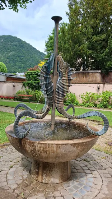 Eine Wiese ist von Wegen durchzogen. Davor wurde eine kreisrunde Fläche mit Kopfsteinpflaster ausgelegt. Darauf steht auf einem runden Sockel eine Schale, in die Wasser von vier aus Kupfer (?) gefertigte, flache Seepferdchen rinnt. Die ca. 3/4m großen Seepferdchen stoßen mit dem Bauch an eine Stange in ihrer Mitte. Der geschwungene Schwanz ist an der Schale befestigt. Hinter einer Mauer im Hintergrund steht ein Baum, auch Häuser sind zu sehen, ein hoher Hügel und der bewölkte Himmel.