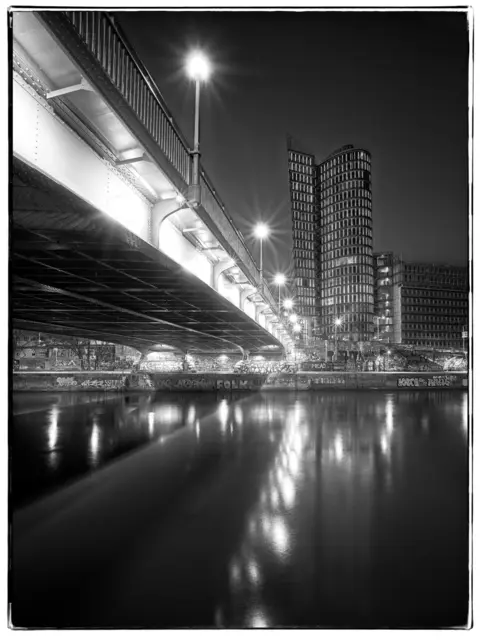 Der Donaukanal zeigt sich hier von seiner stillen, fast grafischen Seite. In Schwarzweiß treten nicht die Farben, sondern Licht, Schatten und Kontraste in den Vordergrund. Die markanten Linien der Brücke führen den Blick zum beleuchteten Uniqa-Tower, dessen Fenster sich im ruhigen Wasser spiegeln. Eine nächtliche Stadtansicht, die die besondere Atmosphäre Wiens zwischen moderner Architektur und urbanem Charme einfängt.