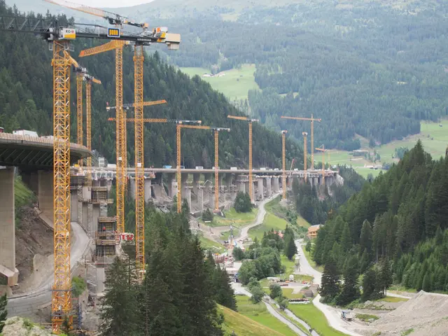 Weitwinkel-Aufnahme der Großbaustelle zum Neubau der Luegbrücke auf der A 13 Brenner Autobahn in Tirol. Im linken Vordergrund stehen dicht gestaffelte, gelbe Turmdrehkrane an einem steilen, grünen Berghang. Die lange Autobahnbrücke zieht sich im Mittelgrund kurvig durch das Wipptal. Im Talboden unter der Brücke verläuft der Flusslauf der jungen Sill, umgeben von Baustraßen, Baufahrzeugen und einem Schotterplatz unter bewölktem Himmel.