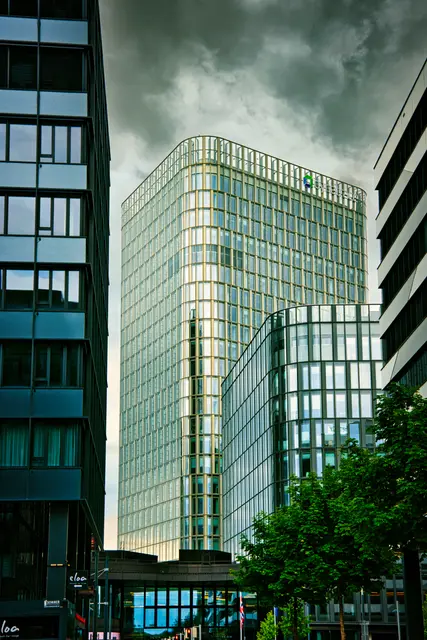 Modernes Hochhaus beim Wiener Hauptbahnhof mit eleganter Glasfassade, eingerahmt von zwei dunkleren Gebäuden. Über dem Gebäude ziehen dramatische Gewitterwolken auf, während die spiegelnden Fenster das Licht einfangen. Im Vordergrund sind Bäume und Teile der modernen Bahnhofsumgebung zu sehen.