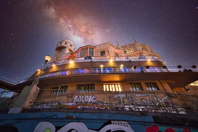 Eine kreativ gestaltete Aufnahme der Wiener Urania bei Nacht aus tiefer Froschperspektive. Im Vordergrund befinden sich bunt besprühte Graffiti-Mauern am Donaukanal. Das beleuchtete Gebäude ragt empor, während im Hintergrund eine nachträglich eingefügte, farbenprächtige Milchstraße am Sternenhimmel leuchtet.