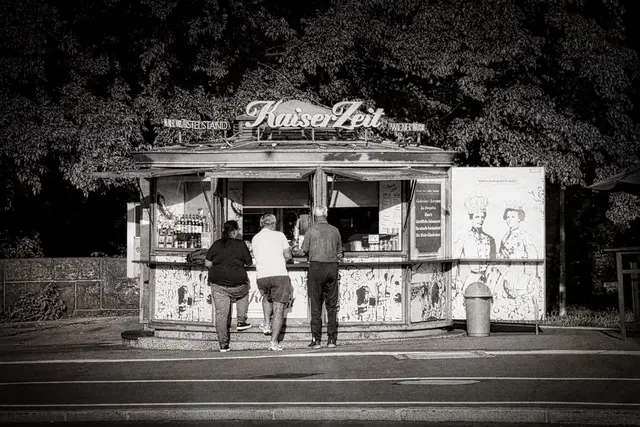 Kontrastreiche Schwarz-Weiß-Fotografie eines traditionellen Wiener Würstelstands namens „KaiserZeit“ am Donaukanal bei der Roßauer Brücke. Drei Personen stehen vor der Theke im Freien und bestellen Essen. Auf der rechten Seite des Kiosks ist eine Illustration des österreichischen Kaiserpaars Franz Joseph und Sisi zu sehen. Im Hintergrund ragen dicht belaubte Bäume empor.
