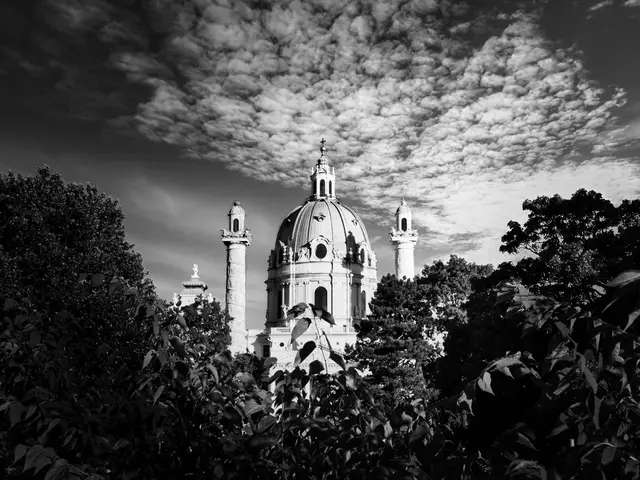 Bild in Schwarz-Weiß mit harten Kontrasten bei tiefer Sonne: Die Kuppel und Säulen der Wiener Karlskirche ragen hinter dunklem Laubwerk hervor, während ein dramatisch strukturierter Wolkenhimmel das Bild nach oben abschließt. Eine zeitlose Komposition, bestimmt von Licht und Schatten.
